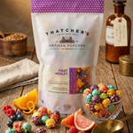 Thatcher's Artisan Popcorn Fruit Medley bag on a wooden table with popcorn and fruits.