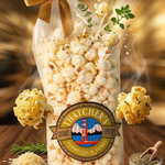 Bag of popcorn with a decorative label on a wooden surface, surrounded by popcorn kernels and herbs.