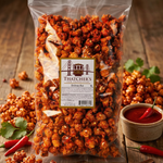 Bag of Thatcher's Sriracha popcorn on a wooden surface with additional popcorn and red peppers.